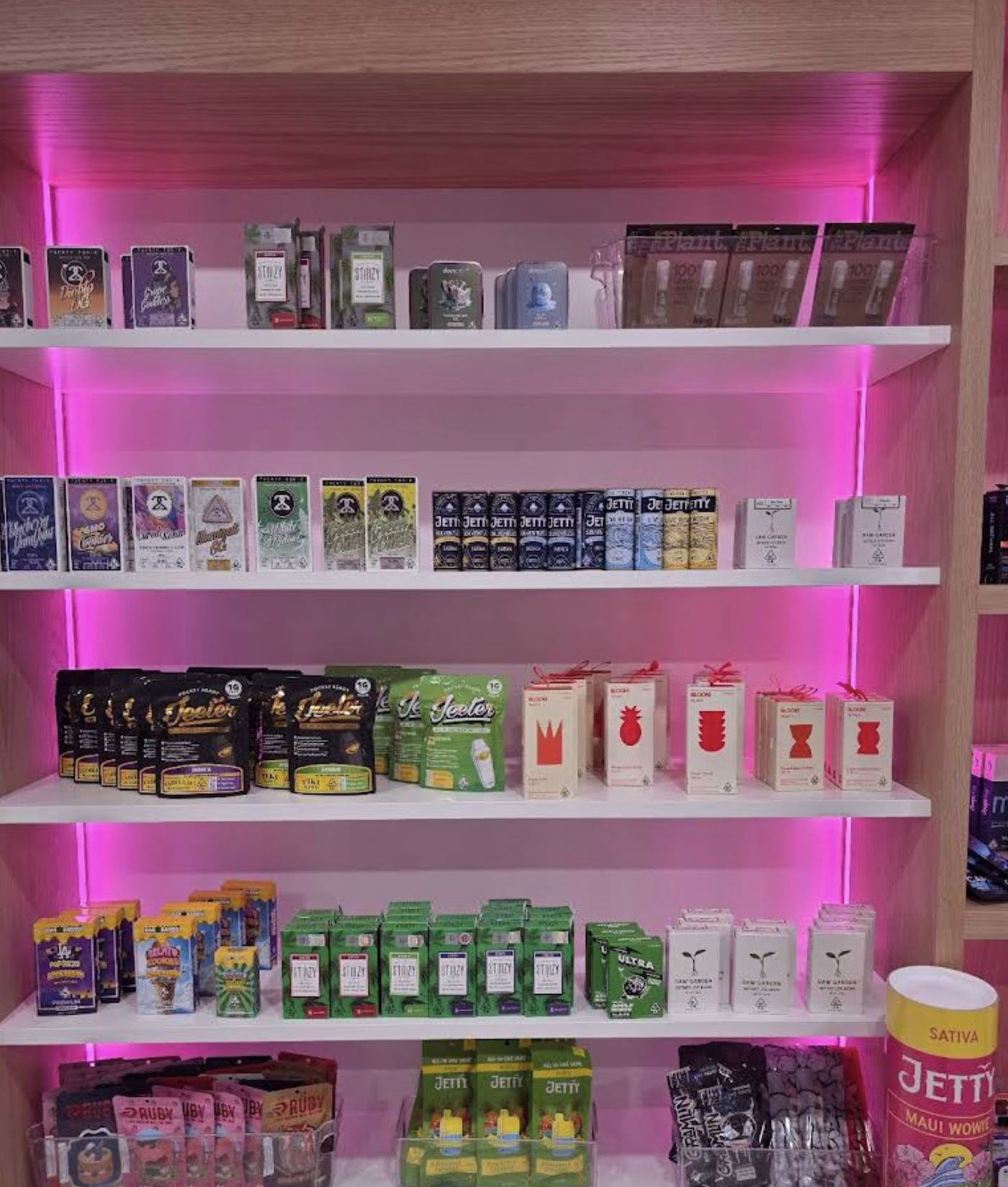 Display shelves featuring various cannabis product packages, including colorful boxes and pouches arranged neatly under pink backlighting